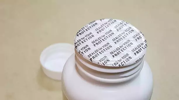 A close-up of a white plastic bottle with a security seal reading "SEALED FOR YOUR PROTECTION" in black text. The bottle cap is placed beside the bottle on a beige surface.