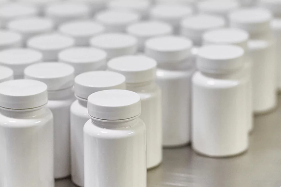 Rows of white plastic bottles with white caps arranged closely together on a flat surface. The bottles are uniform in size and shape, suggesting a mass production setting.