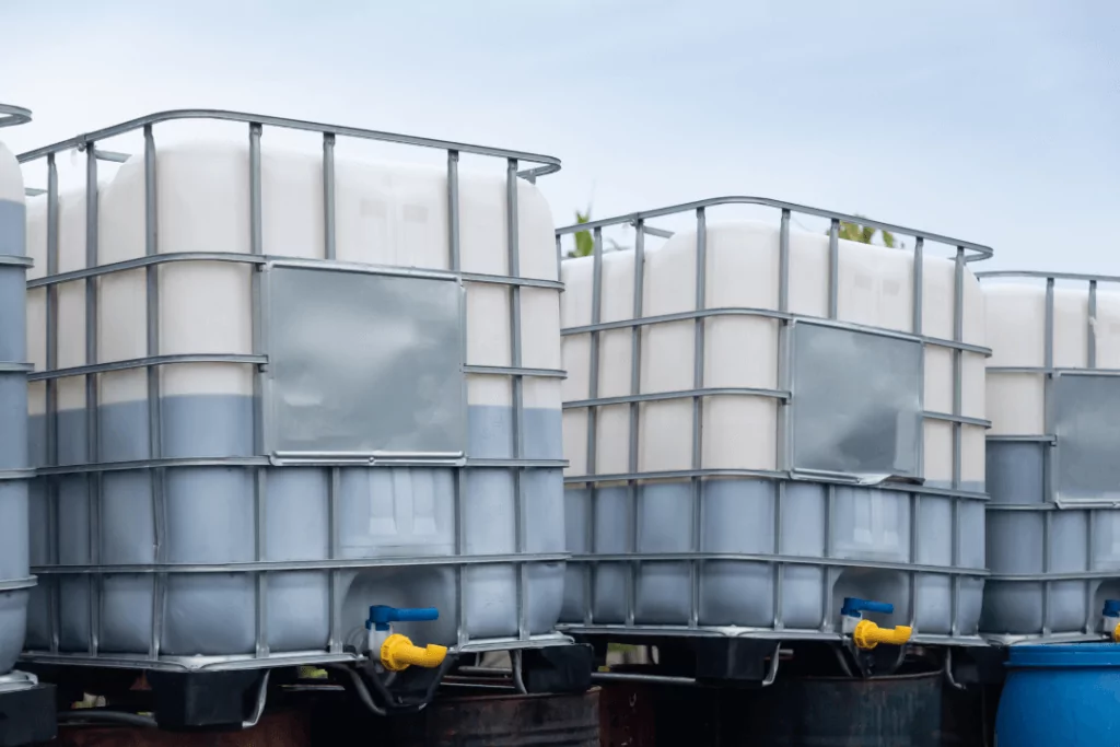 Large industrial containers filled with liquid are stacked outdoors. The containers are white with gray metal cages for support and have blue and yellow spouts at the bottom for dispensing. The sky is overcast in the background.