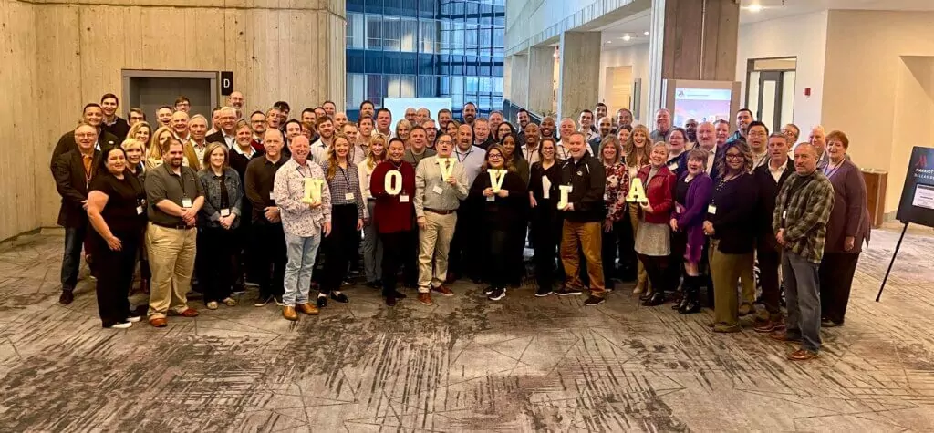 A large group of people smiling and standing closely together in a spacious building with modern architecture. Some individuals are holding large letters that spell 