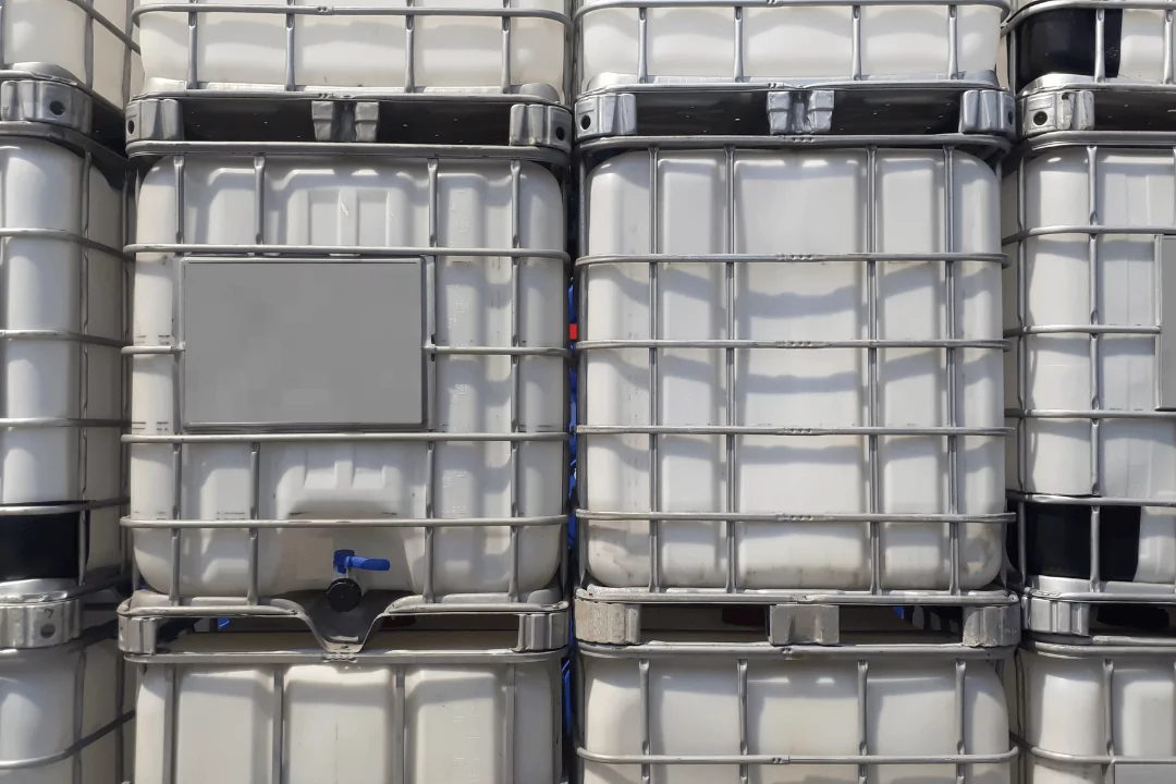 Stacked industrial containers with a grid-like metal frame, used for liquid storage. Each container has a spout at the bottom. The containers are uniform, lined up closely together, and shaded in white and gray tones.