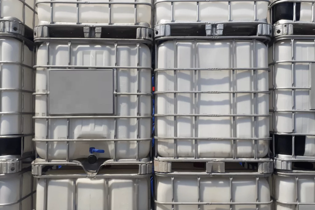 Stacked industrial containers with a grid-like metal frame, used for liquid storage. Each container has a spout at the bottom. The containers are uniform, lined up closely together, and shaded in white and gray tones.
